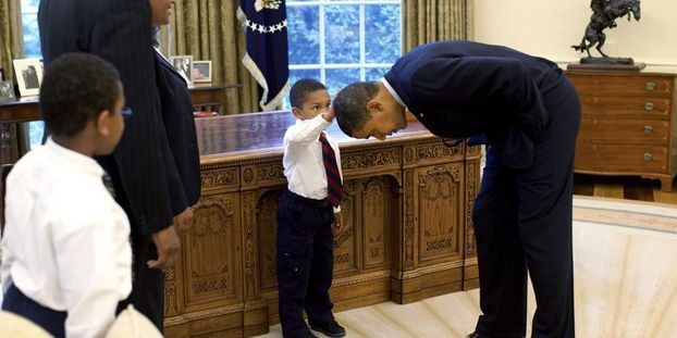 Dans le bureau ovale de la maison blanche, un jeune enfant touche les cheveux du président Barack Obama penché devant lui.