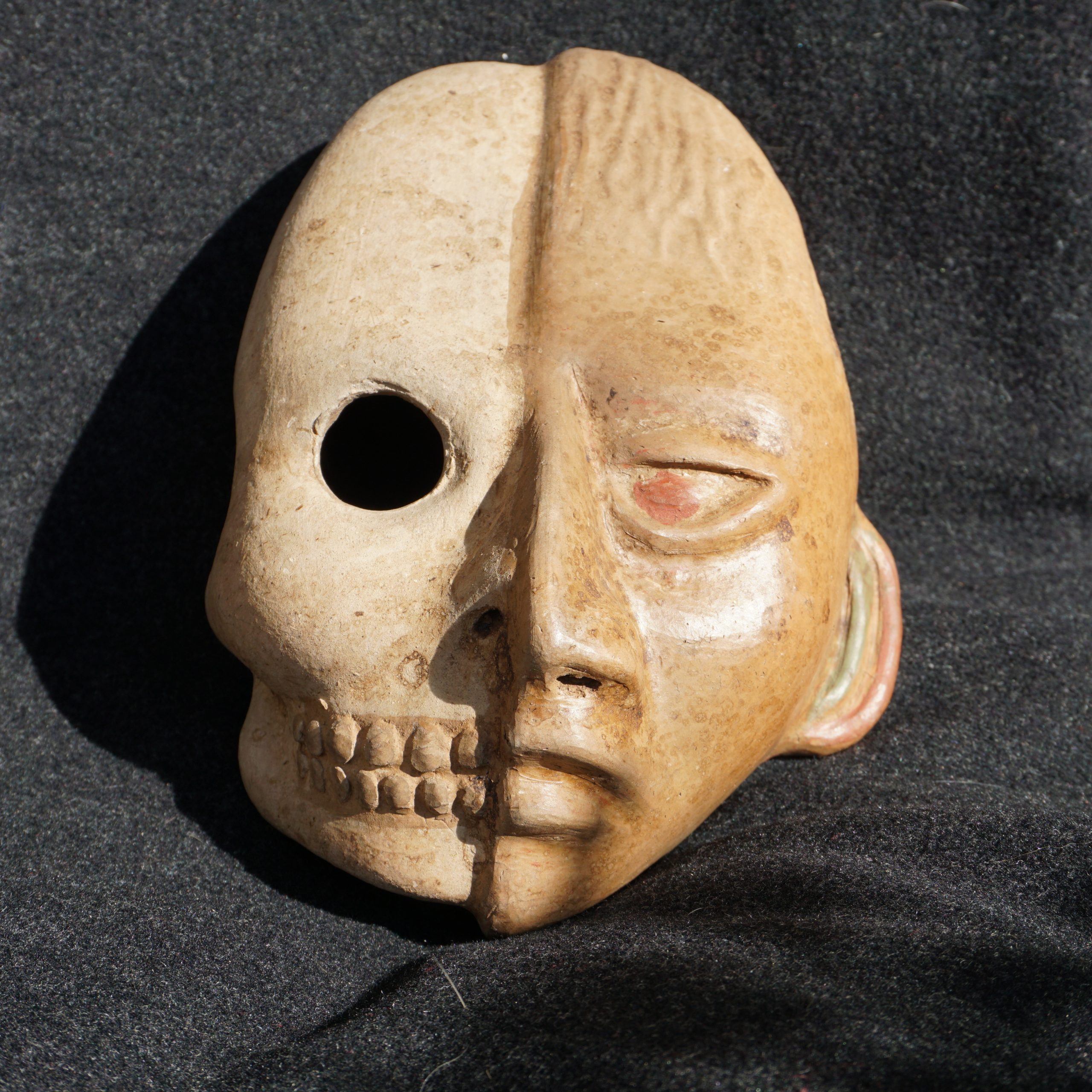 Un masque ramené du Mexique : posé sur un tissu, il représente un visage en pierre scindé en deux dans la verticale, un côté en chair, l'autre en squelette apparent 