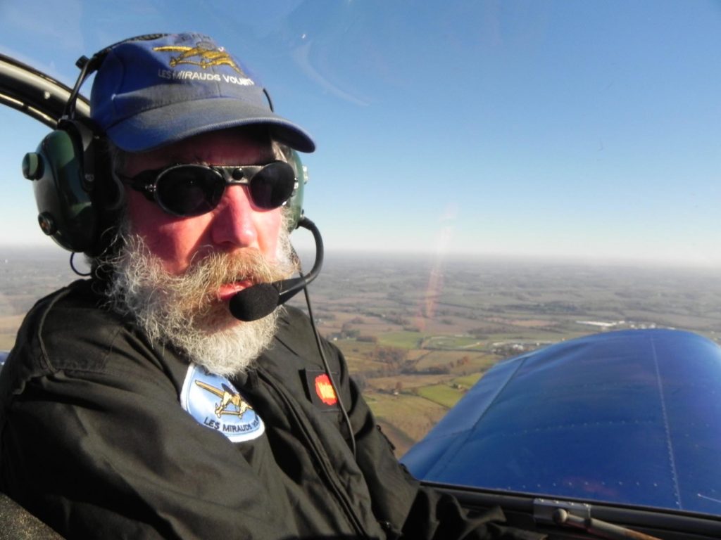 Portrait de Patrice en plein vol, de profil, casquette, combinaison d'aviateur et casque sur un fond de ciel bleu et de champs cultivés.