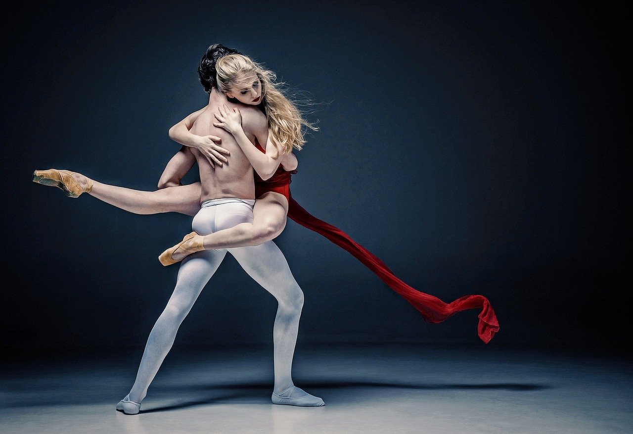 Deux danseurs classiques sur scène lors d'un ballet. Dans un mouvement de porté, l'homme, torse nu et de dos, porte en une étreinte la danseuse qui enroule ses bras et ses jambes en pointes autour de son buste. Les têtes se frôlent tandis que les rubans du costume de la danseuse volent autour d'eux.