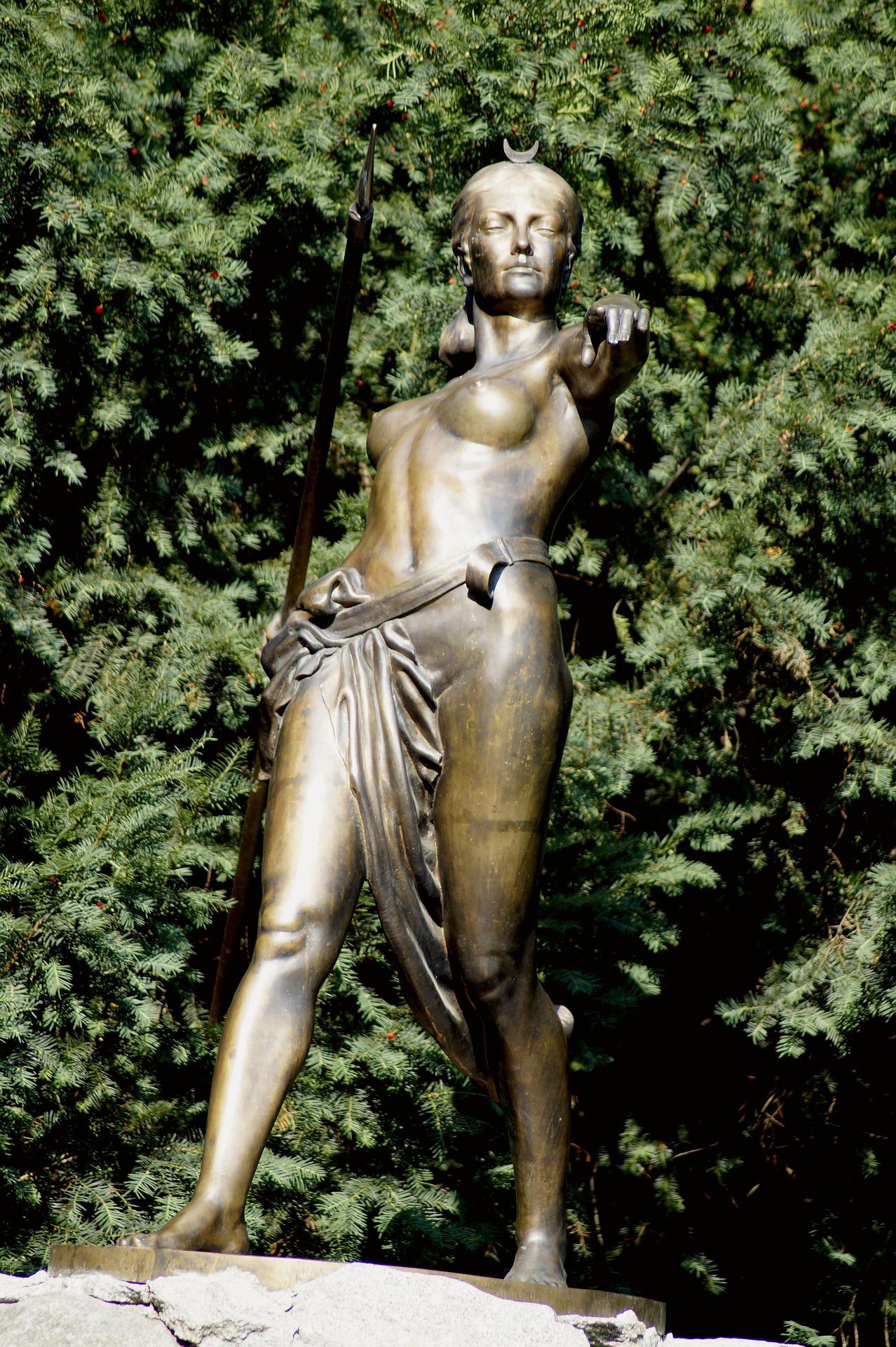 Dans un jardin, statue de bronze de la déesse de la lune, femme chasseuse et guerrière. Nue, de face, un pagne enserrant sa taille, armée d'une lance placée dans son dos, elle pointe du doigt le spectateur en contre-plongée, dans une position de défi. 