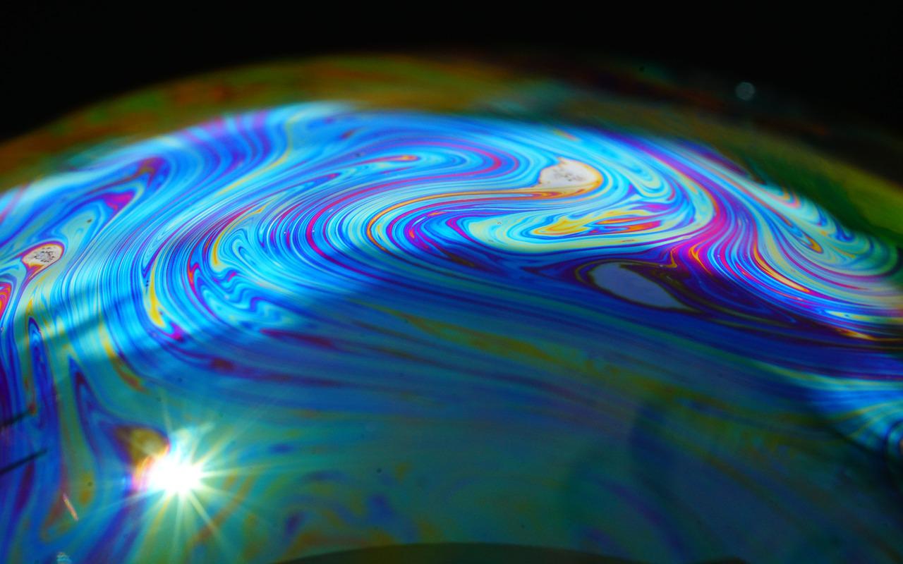 Très gros plan de la courbe d'une bulle de savon en pleine lumière: des volutes multicolores se déploient le long de la surface, tandis qu'un point renvoie le soleil en une étincelle.