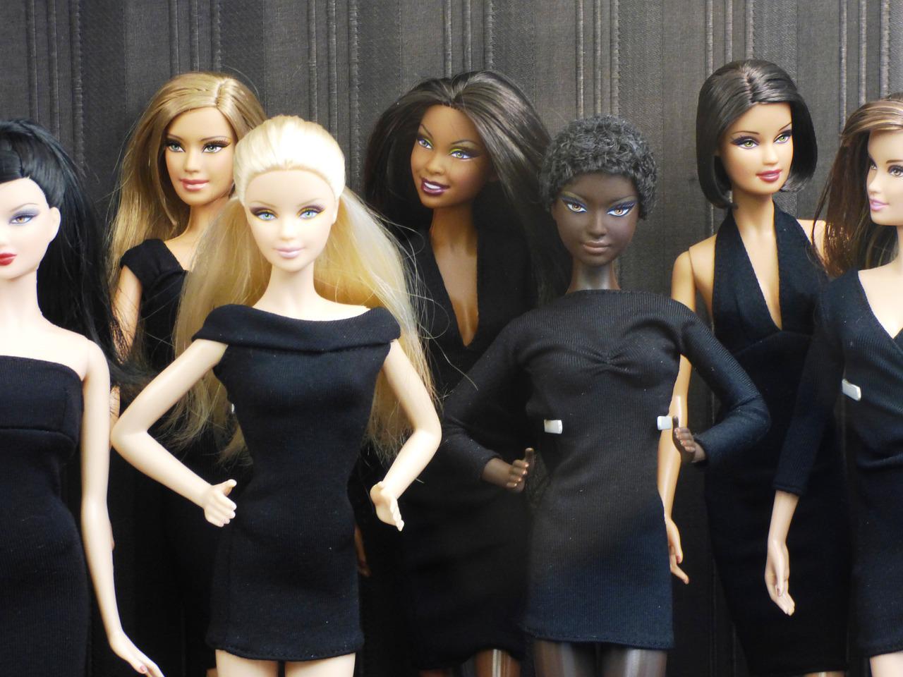Sept poupées Barbie disposées comme posant devant la caméra. Brunes, blondes, cheveux crépus, asiatique, métis, blanches ou à la peau noire, elles se tiennent droites et immobiles, les mains sur les hanches ou derrière le dos.