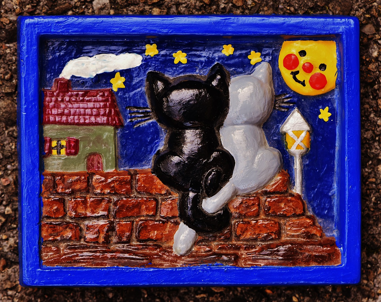 Moulage en plâtre plat dont les détails de l'image apparaissent en relief: l'illustration représente deux chats de dos posés sur un mur, les queues enlacées, face à la lune et à une maisonnette. Le style et la peinture sont enfantins.