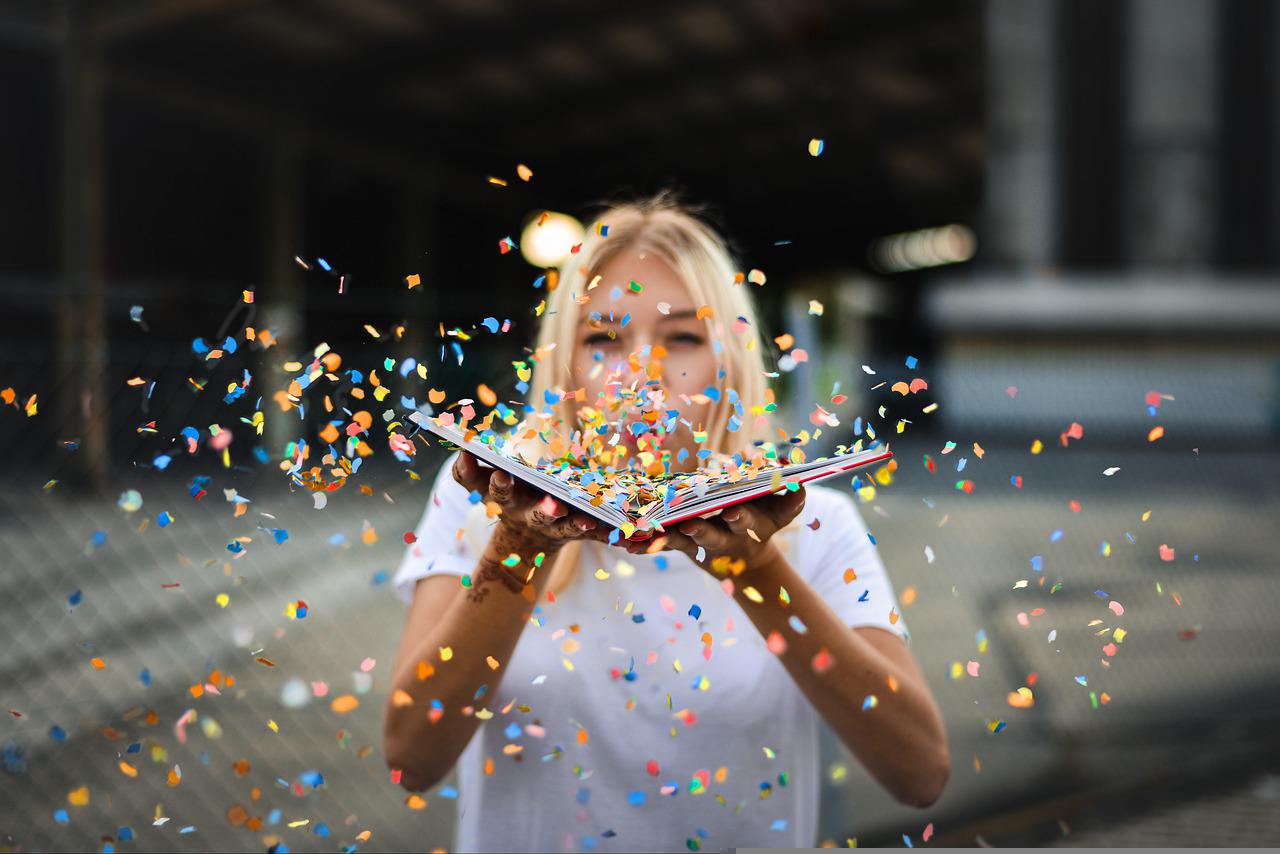 De face, une jeune femme souffle une multitude de confettis depuis un livre tenu ouvert devant sa bouche. Les petits fragments de papiers colorés envahissent la photographie.