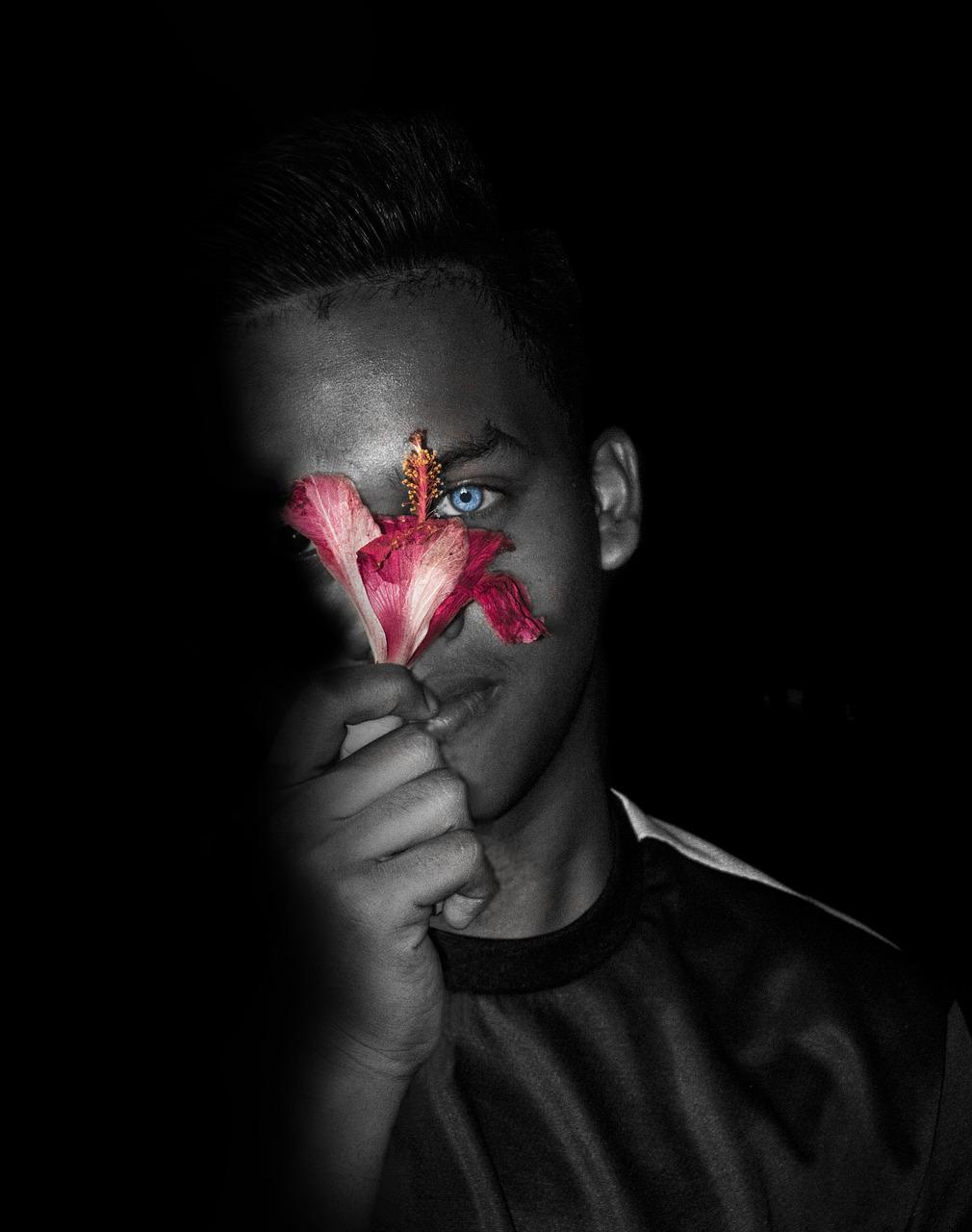 Portrait en noir et blanc d'un jeune homme dans l'obscurité qui tient contre une moitié de son visage une fleur. Le pistil et les pétales épousent d'une caresse les courbes de son visage. Seuls la fleur et l'oeil de l'homme sont en couleur.