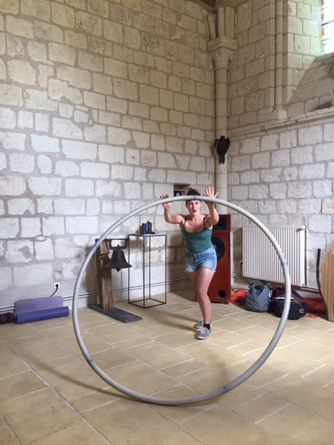 Une démonstration d'acrobatie dans une salle aux hauts murs de pierre: une femme manie un cerceau d'un diamètre plus grand qu'elle en le faisant rouler sur le sol.