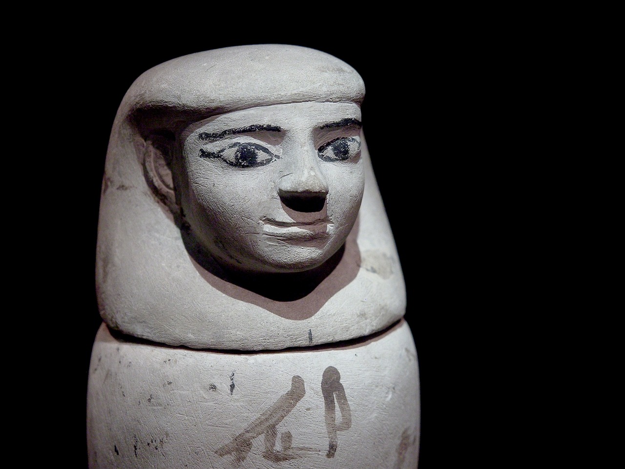 Gros plan sur la tête d'un vase canope égyptien. En terre cuite, il représente une tête d'égyptien souriant, à la coiffe et  au maquillage d'époque.