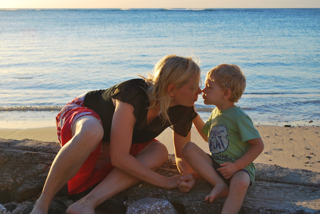 Baiser esquimau entre une mère et son enfant, assis sur la jetée en pierre le long d'une plage, avec la mer en fond. Les nez se touchent, la mère se penche vers l'enfant qui l'attrape par l'épaule.