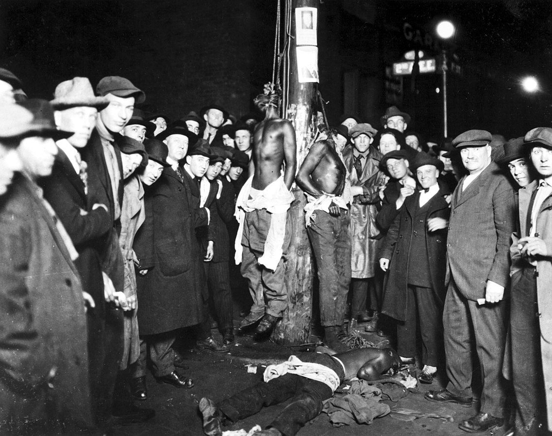 Photographie en noir et blanc du lynchage de Duluth. Une foule nombreuse d'hommes blancs rassemblés autour d'un poteau auquel deux hommes noirs torses nus sont pendus. Un troisième homme noir gît à terre.