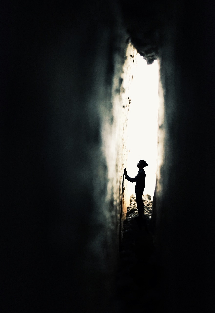 Photographie prise à travers une crevasse  dans un mur, formant comme une meurtrière. A travers celle-ci, on distingue sur un fond de lumière, la silhouette d'un homme, de profil, qui pose ses mains sur le mur devant lui. Tout est plongé dans le noir, sauf cette scène perçue à travers la fissure.