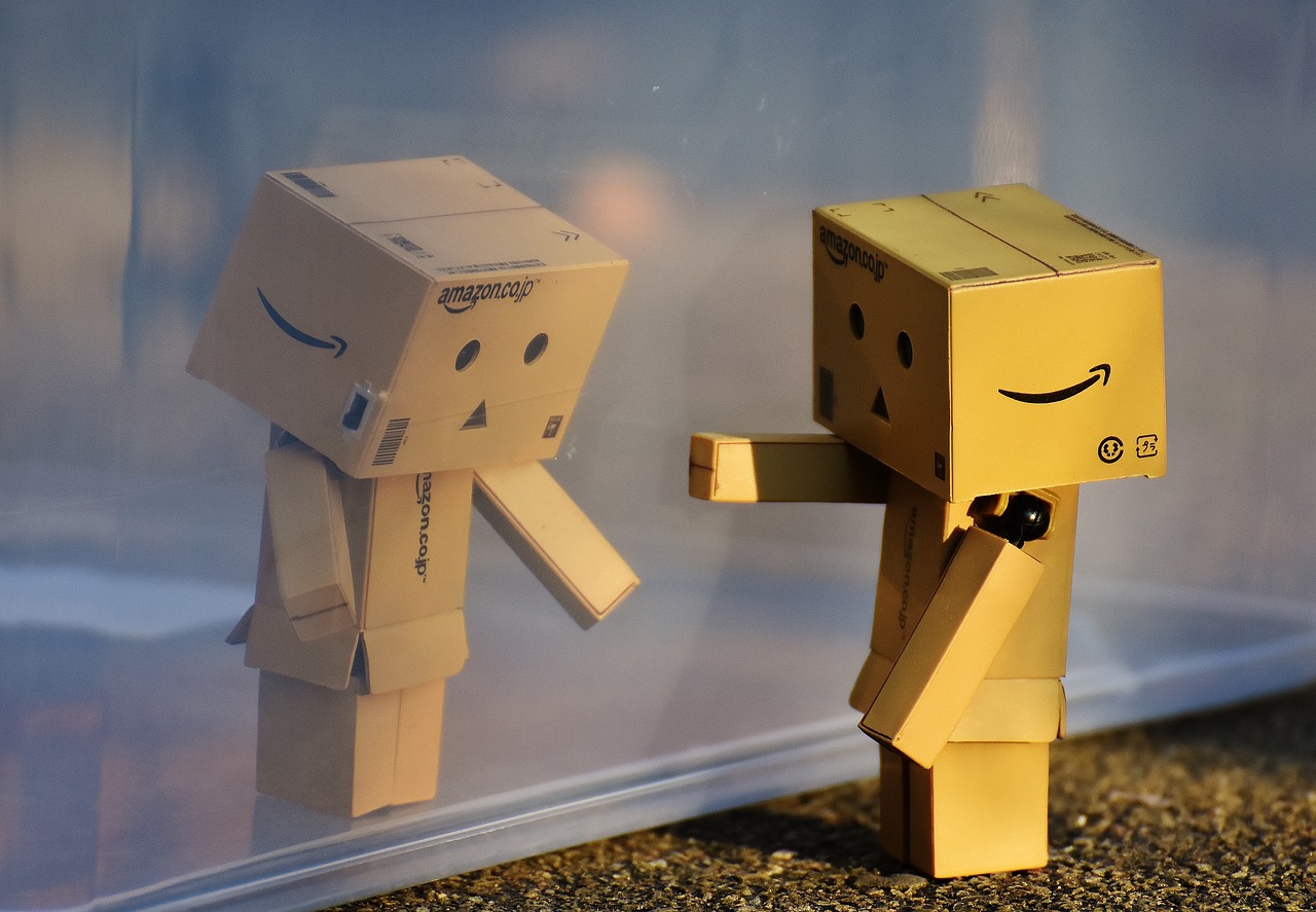 Deux petits bonshommes de style Lego, composés de cartons de livraisons (un colis pour la tête, un pour le corps et pour chaque membre avec logo Amazon) se font face à travers une vitre. Leurs visages composés de deux yeux et une bouche dessinés expriment l'étonnement de ne pas pouvoir se toucher.