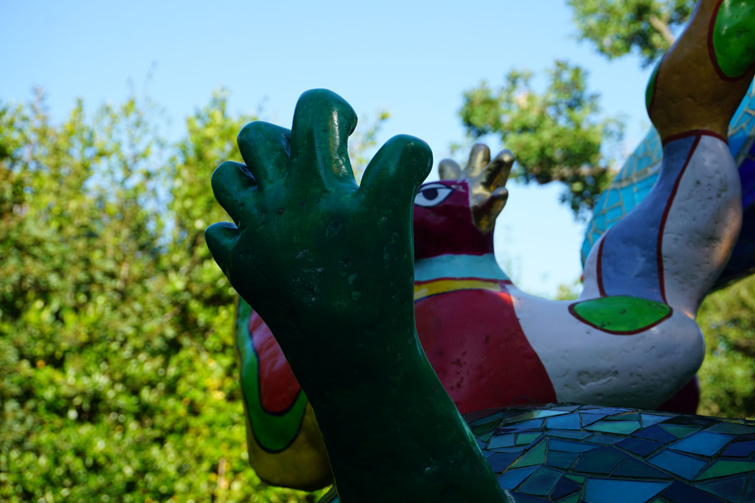 Une partie d'une des sculptures du jardin, vue en contre-plongée. Une main de couleur verte, les doigts légèrement repliés comme pour attraper quelque chose se dresse, plantée dans le socle. Derrière, on distingue des éléments colorés du reste de la statut et une haie au loin.