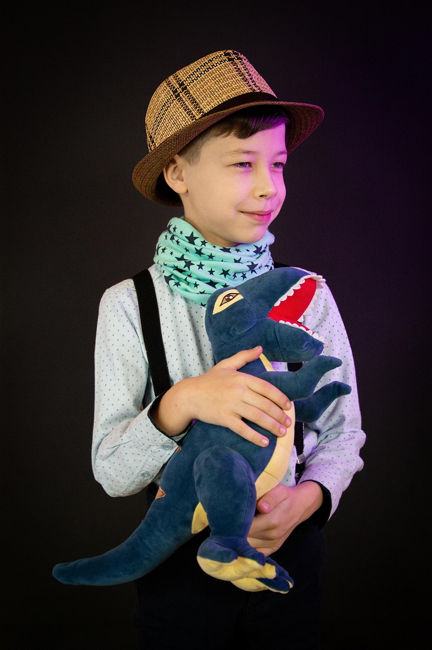 Sur fond uni, un garçon pose fièrement en tenant contre lui un T-Rex en peluche. Le garçon se tient de face, souriant, la tête légèrement de profil, le regard porté vers un hors champ à droite. Il porte une chemise, des bretelles, une écharpe et un chapeau dans un style un peu rétro. Il tient le dinosaure en passant une main entre ses pattes, l'autre posée délicatement sur son dos.