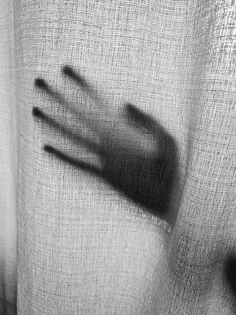 Derrière un rideau en lin blanc, l'ombre d'une main apparaît, comme si elle se posait délicatement sur le rideau sans le toucher totalement, les doigts un peu étirés par le reflet, presque comme une empreinte à l'encre sur le tissu.