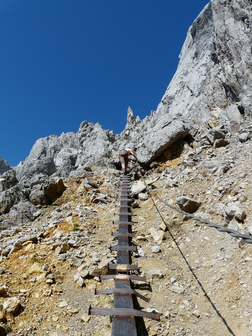 Vue en contre-plongée d'une via ferrata. Le spectateur se trouve en contre-bas, comme s'il était en train de monter le long de la voie composée d'une barre de fer ancrée dans la roche et surmontée, à espaces égaux, de chevons (principe de la voie ferrée). A droite de la voie, la ligne de fer appelée "ligne de vie" qui monte jusqu'en haut. Tout en haut, la vue voie se perd dans les roches pointues et un grimpeur, que l'on distingue à peine, est en train de monter, non pas face à la voie, mais en biais, face à la ligne de vie à laquelle il se tient, légèrement penché.