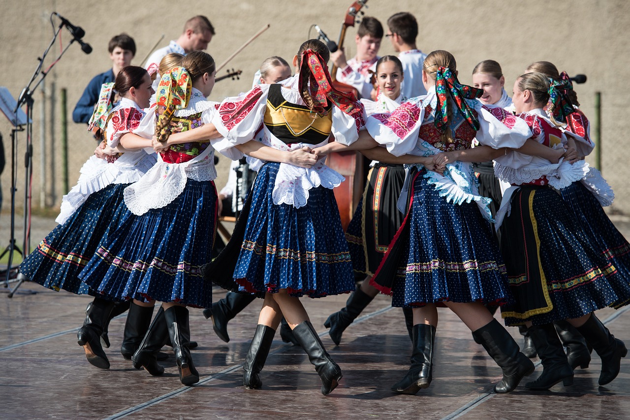 Danse en cercle folklorique de l'Europe de l'ouest. Derrière, un groupe de musiciens à cordes, devant, un cercles de jeunes femmes en bottes, jupons à carreaux, chemises bouffantes, gilets en crochet et longs rubans dans les cheveux. Elles tournent en se tenant par la taille.
