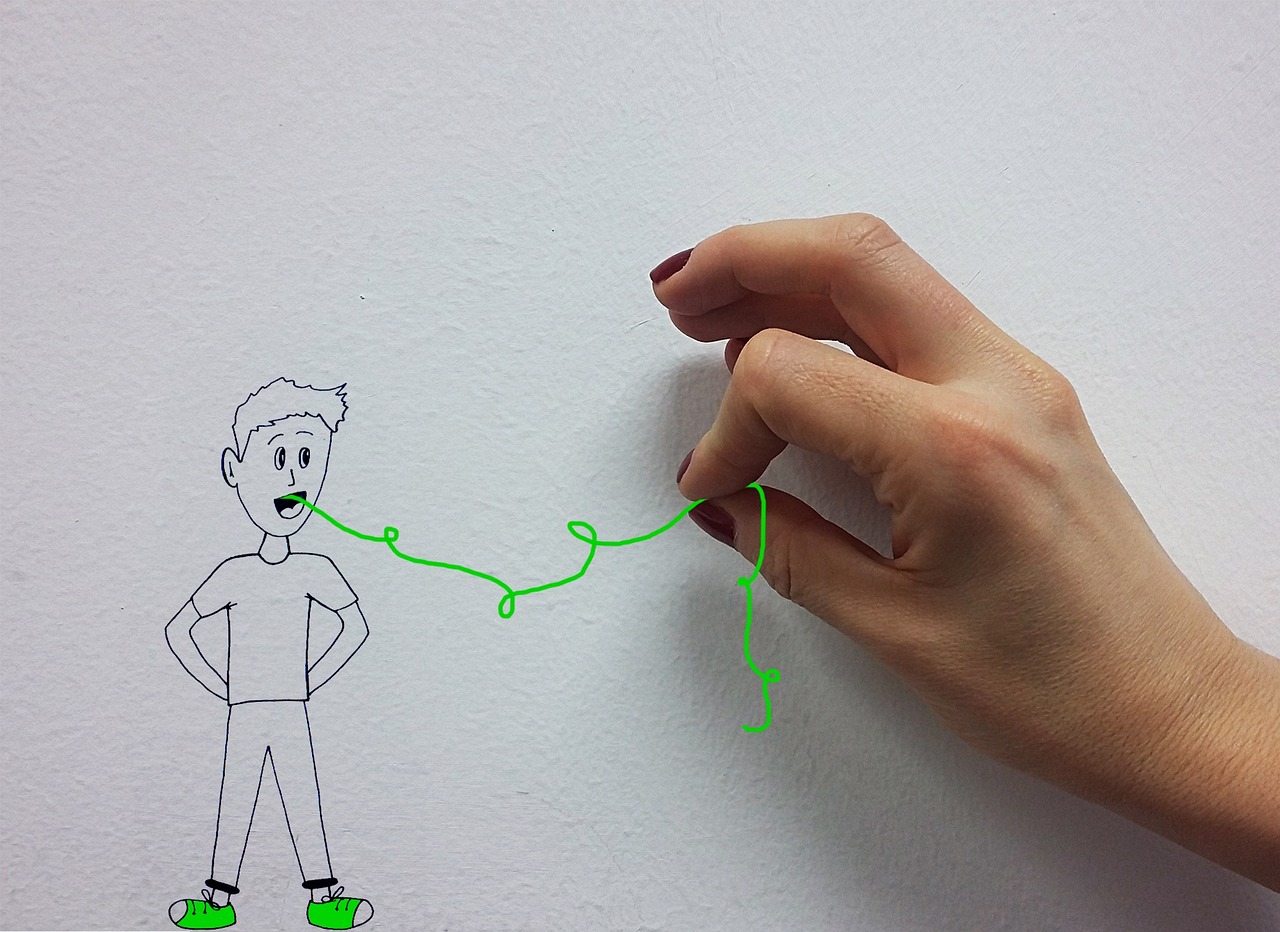 Sur une feuille de papier blanc, un petit bonhomme est dessiné. C'est un garçon habillé d'un t shirt, d'un pantalon et de baskets. Une main humaine s'approche du dessin et tire un fil imaginaire qui sort de la bouche du garçon dessiné en train de parler. Le fil est dessiné au feutre vert fluorescent.