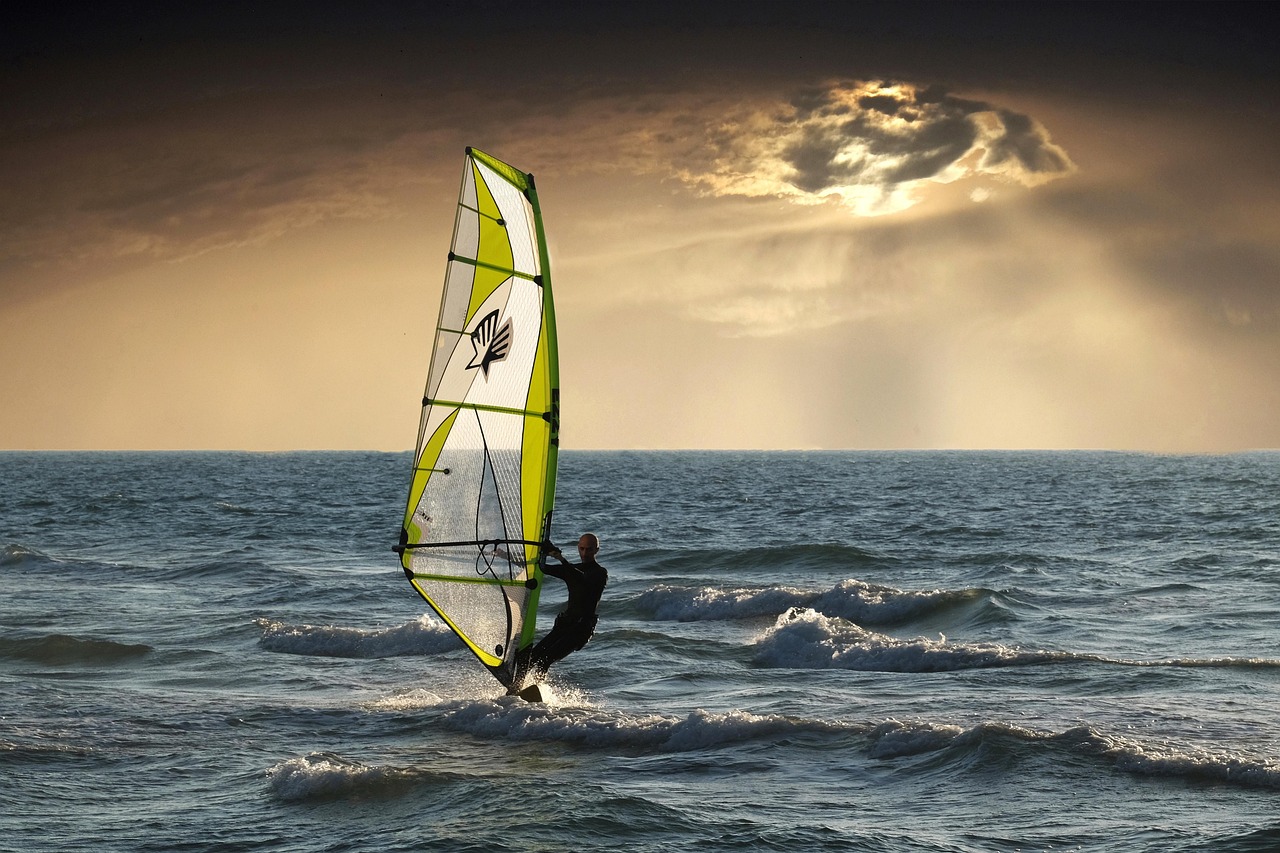 Sous un ciel de fin de journée dont la lumière traverse un nuage, un homme, de face, fait de la planche à voile, seul au milieu de l'eau.