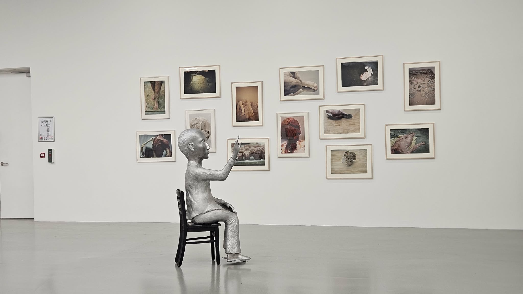 Dans une galerie de musée, une sculpture au centre et des photographies encadrées au mur du fond - disposées en quinconce. La sculpture, au centre de la pièce, est un bonhomme, tout argenté, presque à taille humaine, chauve et en costume trois pièces, assis de profil sur une chaise noire. Il tend la main devant lui comme s'il voulait taper dans la main du visiteur ou comme s'il le stoppait. Avec la perspective de la photo, on dirait qu'il touche les tableaux.