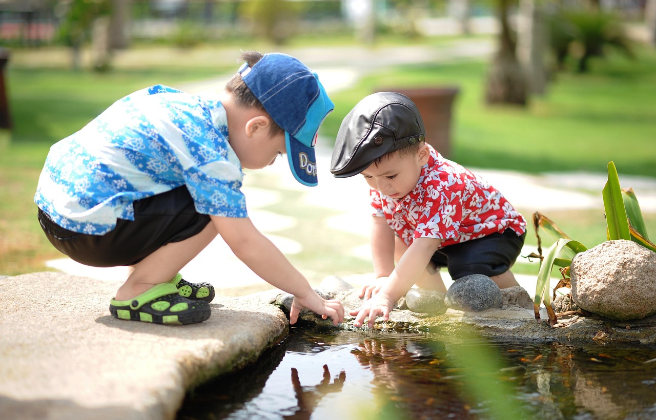 Dans un jardin public, deux petits garçons sont accroupis sur le chemin de pierres qui borde une mare. Ils tendent tous les deux la main vers quelque chose qu'ils essaient soit de toucher soit d'attraper à la surface ou dans l'eau. 