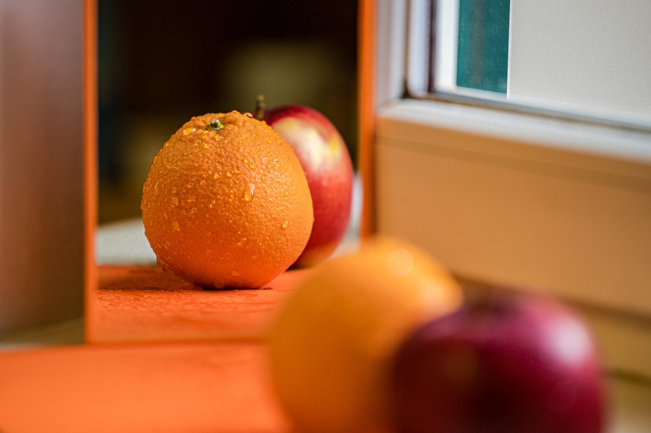 Sur une table, une orange est posée devant une pomme rouge face à un miroir. Dans le miroir, se reflètent l'orange au premier plan - dont les aspérités de la peau sont mises en valeur par des gouttelettes d'eau comme de la rosée -, au second plan, la pomme - dont la peau lisse est accentuée par le reflet du soleil. Les deux fruits sont disposés en quinconce.