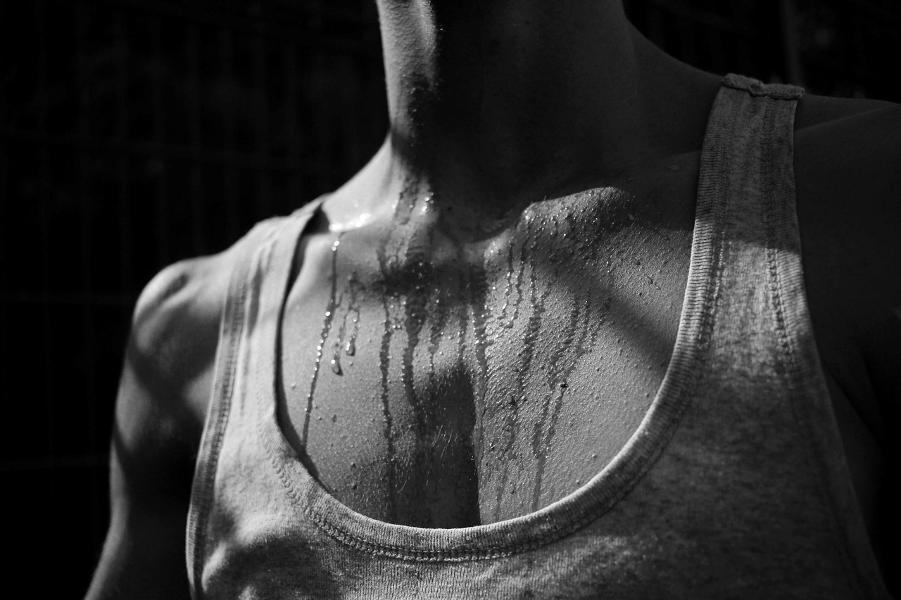Gros plan en noir et blanc de coulées de transpiration sur le buste d'un humain. La tête et la poitrine sortent du cadre donc on ne connaît pas le sexe de la personne. L'individu porte un débardeur de type marcel; les coulées partent de son cou pour descendre sous l'encolure. 