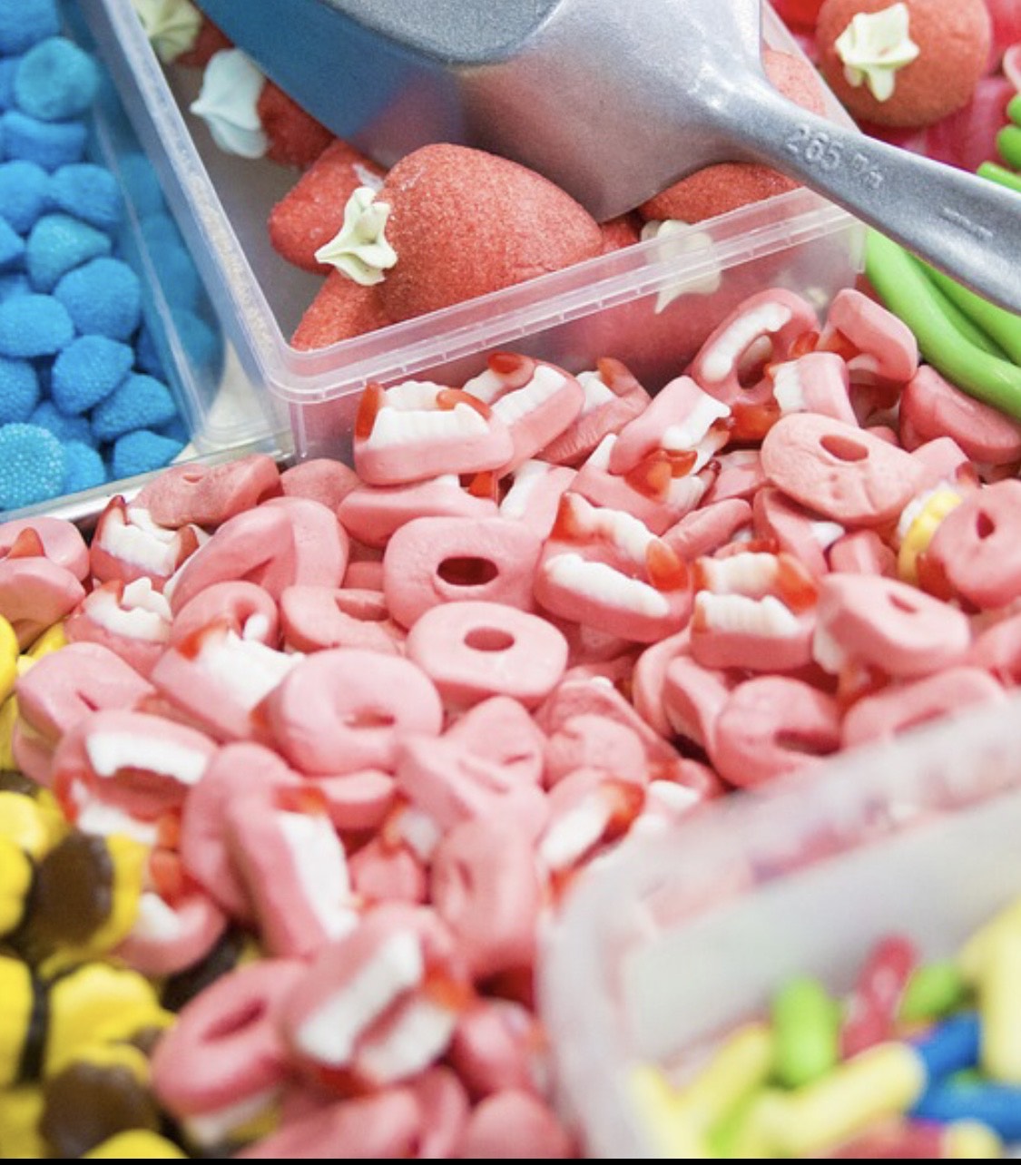 En gros plan, un étal de bonbons. Plusieurs boîtes transparentes sans couvercle se côtoient: des petites boules bleues en haut, des flans caramels en bas, de grosses fraises au sucre sur lesquelles repose la petite pelle pour se servir. Au centre, qui prend le plus de place sur l'image, des dentiers de vampire à la fraise (rose pour les gencives, blanc pour les dents, rouge pour les canines).  