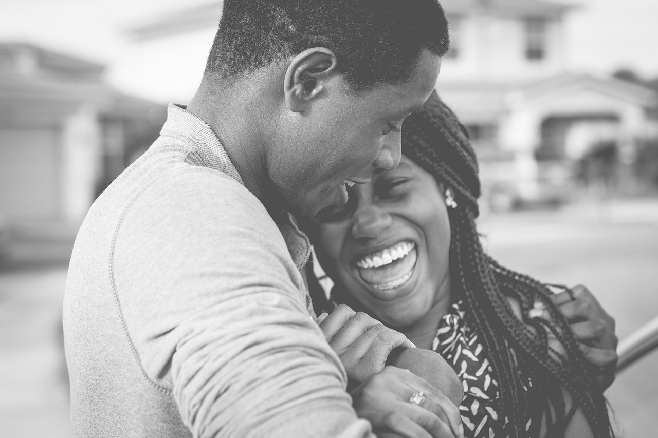 Portrait en noir et blanc d'un couple qui se fait un câlin. La femme, à droite, dans un grand éclat de rire, pose sa tête sur le torse de l'homme à gauche, qui, lui, souriant, incline la sienne vers elle. Sa main gauche enserre l'épaule de la femme qui elle agrippe le poignet droit de l'homme, au niveau de leur poitrine. Ils ont la trentaine, la peau noire, l'homme a des cheveux courts, la femme a de longues tresses qui glissent entre leurs doigts.