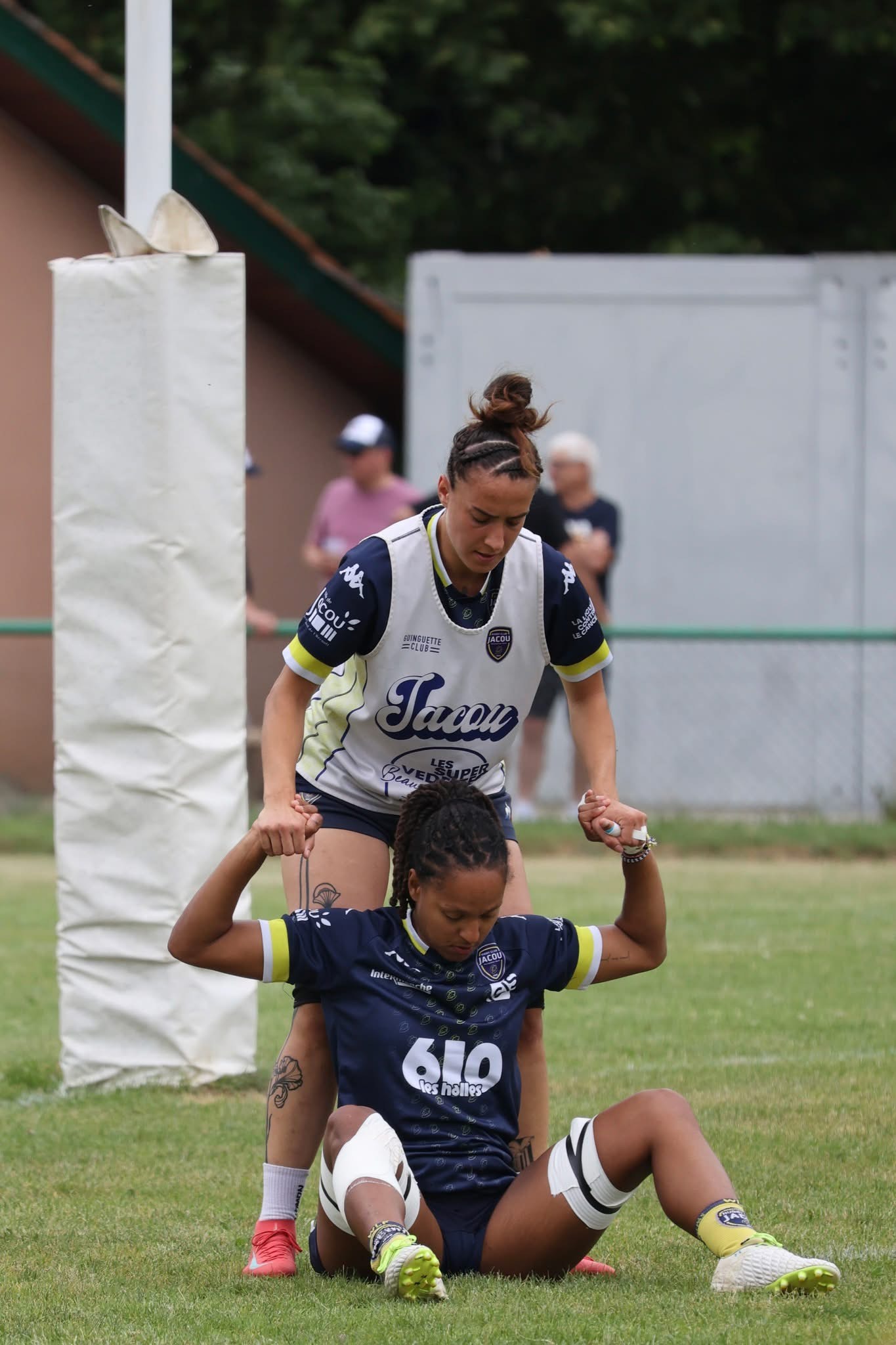 Deux joueuses de rugby en train de s'échauffer mutuellement avant un match. L'une est assise, les jambes ancrées dans le sol, l'autre est debout derrière elle. Celle qui est assise colle son dos contre les jambes de celle debout derrière. La joueuse assise a les bras en l'air au-dessus de sa tête pour donner les mains à celle debout. Celle de derrière assure une poussée vers le bas dans les mains de l'autre joueuse et dans les jambes contre le dos. En poussée contraire, la joueuse assise pousse avec les mains vers le haut et en arrière avec son dos contre les jambes de la seconde.