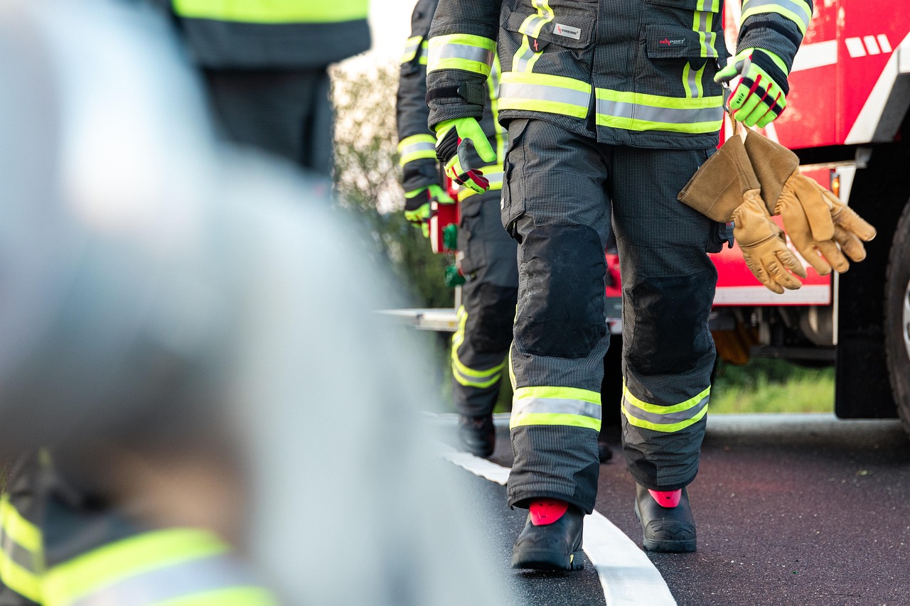 Des pompiers se déplaçant en ligne droite, les uns derrière les autres, entre deux de leurs camions sur une route goudronnée. On ne voit que jusqu'à leur buste. Ils sont en tenue complète, les mains recouvertes de gants et ont à leur ceinture d'autres sortes de gants.