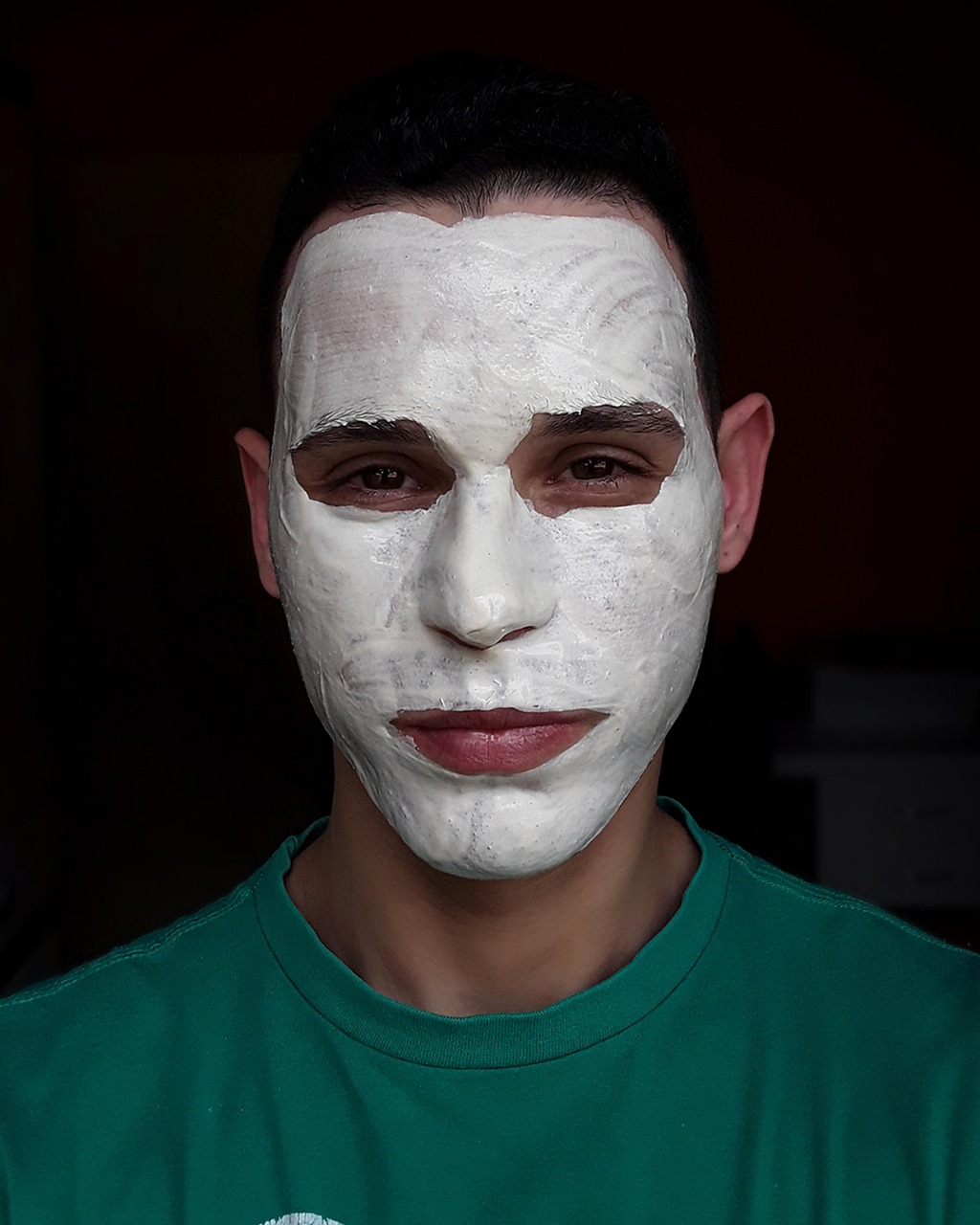 Portrait, de face, d'un homme en t-shirt dont le visage est recouvert d'un masque d'argile blanc. Seuls ses yeux, ses oreilles, sa bouche et son cou sont encore nus.