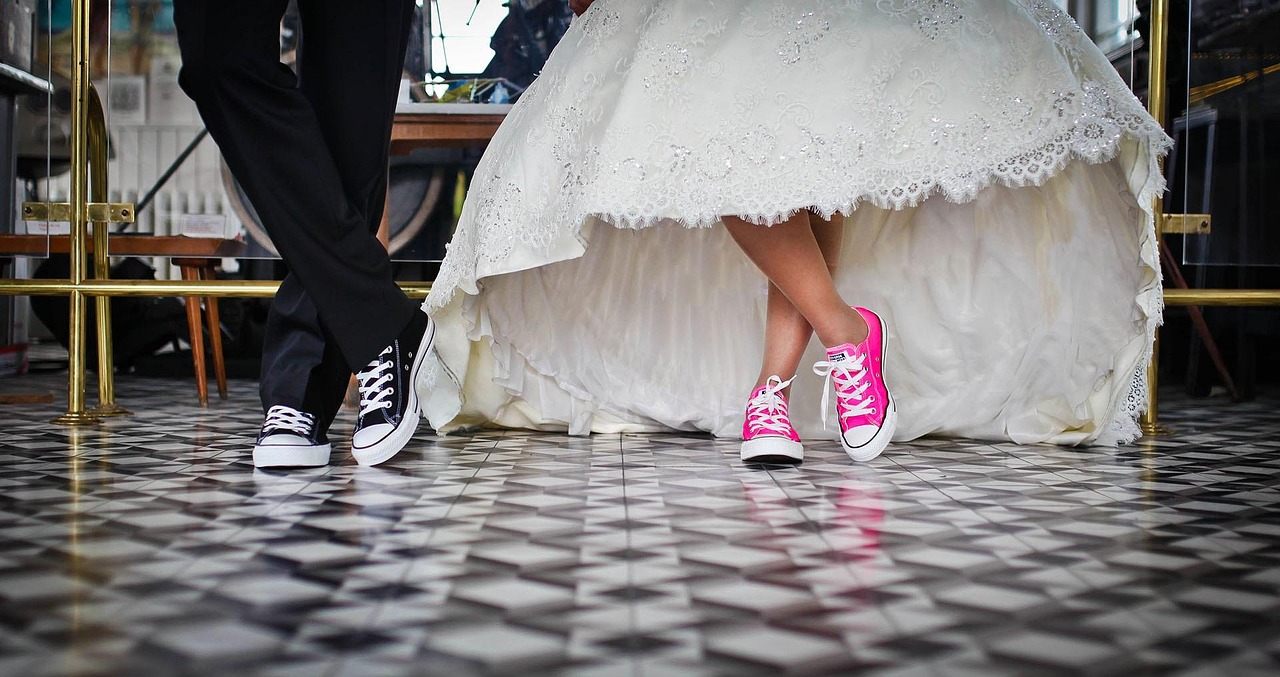 Un couple de mariés en habits classiques de mariage, dans une église ou à la mairie (sol carrelé à damiers, vitrines et dorures en fond), qu'on ne voit que jusqu'aux genoux. À gauche, le marié porte un bas de smoking noir, à droite, la mariée porte une robe en large corolle de dentelle très travaillée, plus courte devant et trainante derrière. Moins classiquement, tous deux portent des converses, noires pour l'homme et rose bonbon pour la femme.